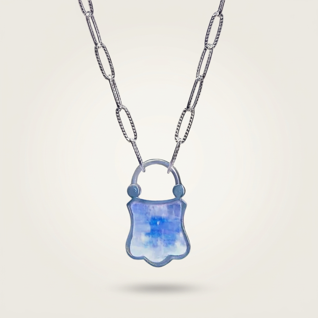 A silver paperclip chain necklace with a shield-shaped pendant featuring a blue and white marbled pattern, set against a light beige background.